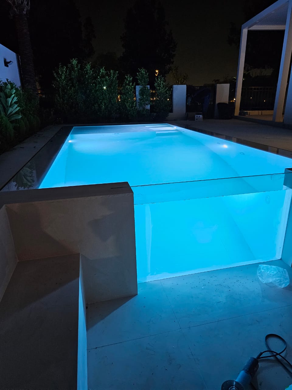 Pool illuminated with blue light at night, surrounded by plants and a building.